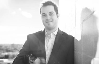 Photo monochrome de Daniel Gervais qui tient un violon, près d'un mur, souriant à la caméra.