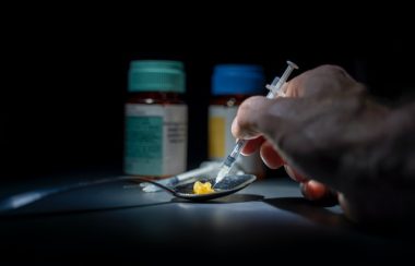 A hand holds a syringe to a spoon with two pills in it. There are two pill bottles in the background.