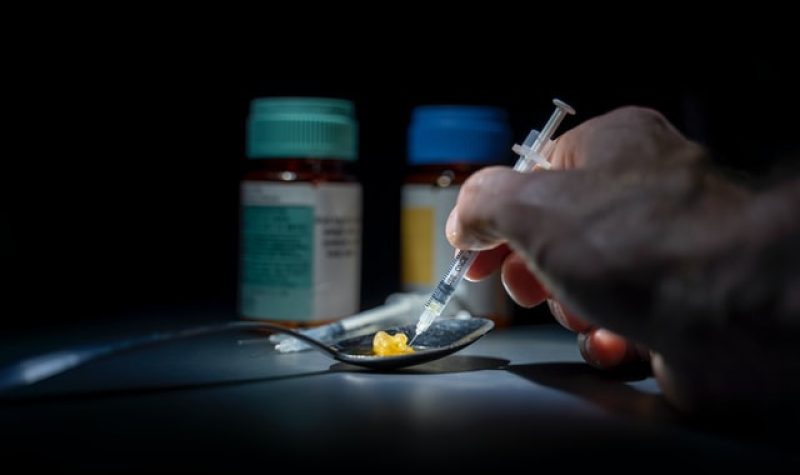 A hand holds a syringe to a spoon with two pills in it. There are two pill bottles in the background.