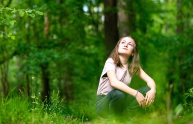 Teenage girl squats down in a forest and pensively looks up at the treetops