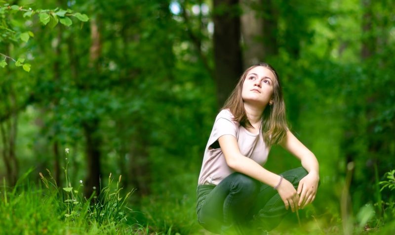 Teenage girl squats down in a forest and pensively looks up at the treetops