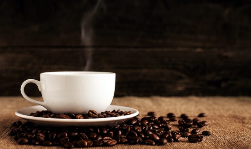 A white cup and saucer with steam coming off and coffee beans scattered around on the flat surface.