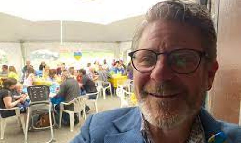MLA Bruce standing at an event in front of a group of people eating lunch wearing a blue jacket.