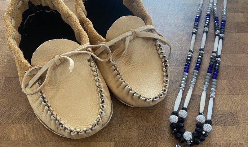 A beige pair of leather moccasin's are displayed alongside a beaded black,blue and white necklace with an arrowhead at the center of the necklace.