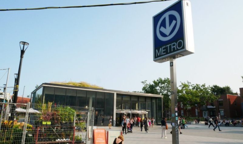 The entrance to a metro station in Montreal is fronted by a large sign with a down-pointed arrow, and surrounded by greenery.