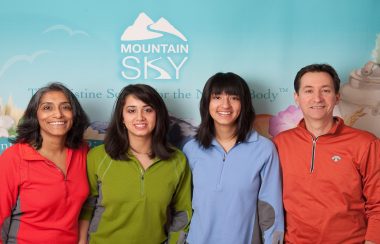 Nina their two daughter and Raynald stand in front of light blue backdrop with the mountain sky logo on it.