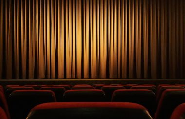 Vu d'une personne assise dans un théâtre vide avec les sièges rouges devant lui, les rideaux fermés et une lueur de lumière sur le rideau jaunâtre.