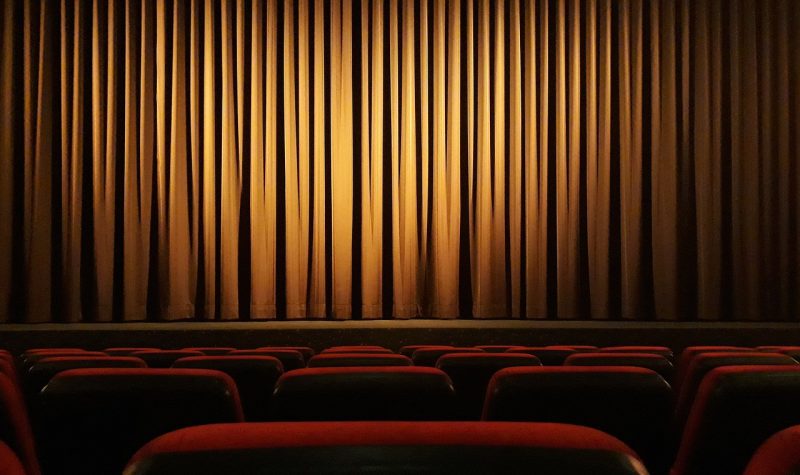 Vu d'une personne assise dans un théâtre vide avec les sièges rouges devant lui, les rideaux fermés et une lueur de lumière sur le rideau jaunâtre.