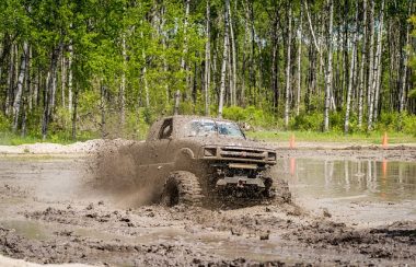 Un camion deux portes avec de grosses roues est dans un trou de boue. Il est complètement couvert de terre. Derrière le trou de boue, une grande forêt.