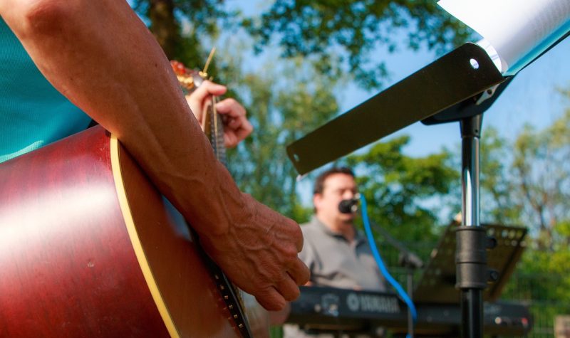 A person's hands playing guitar with an easel and paper in front of the person, A man can be seen in the background playing a keyboard. The two people are performing in an outside venue with trees lining the background.