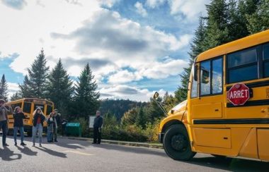 À gauche, des personnes photographient l'autobus jaune qui se trouve à droite. La toile de fond est composée d'arbres sous un un ciel nuageux avec quelques éclaircies.