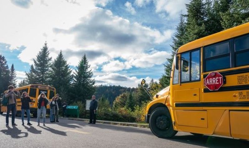 À gauche, des personnes photographient l'autobus jaune qui se trouve à droite. La toile de fond est composée d'arbres sous un un ciel nuageux avec quelques éclaircies.