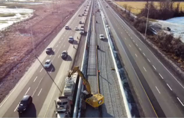 A highway is seen from an aerial view. There are cars driving in the direction of the camera on the left side; the right side is empty. The road is bordered by grassy areas on either side. In the middle of the highway, construction can be seen, with a crane lifting sections of rail and setting them on a track.