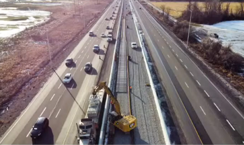 A highway is seen from an aerial view. There are cars driving in the direction of the camera on the left side; the right side is empty. The road is bordered by grassy areas on either side. In the middle of the highway, construction can be seen, with a crane lifting sections of rail and setting them on a track.