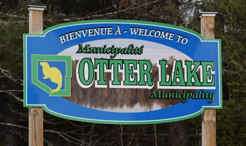 The sign for Otter Lake Quebec, blue with green letters and a yellow otter silhouette.