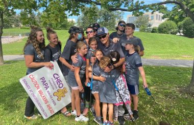 An elderly man wearing dark glasses and carrying walking poles is embraced by a large group of children and young people in a grassy area.