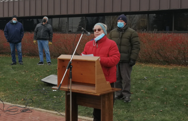 A woman in a red winter coat stands behind a wooden podium outside of a building on an overcast day with three people behind her in masks