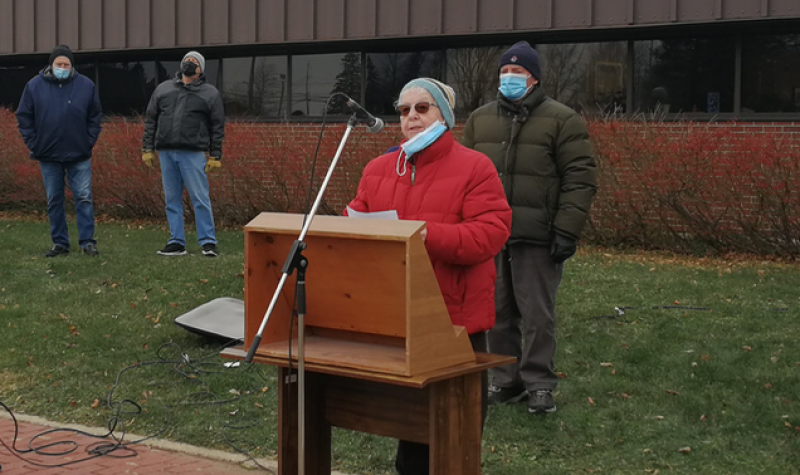 A woman in a red winter coat stands behind a wooden podium outside of a building on an overcast day with three people behind her in masks