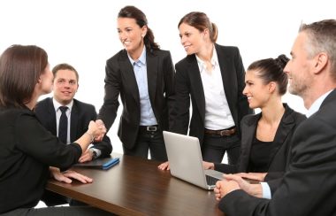 Six personnes vêtues en habit d'affaires autour d'une table avec ordinateur portable et une poignée de main
