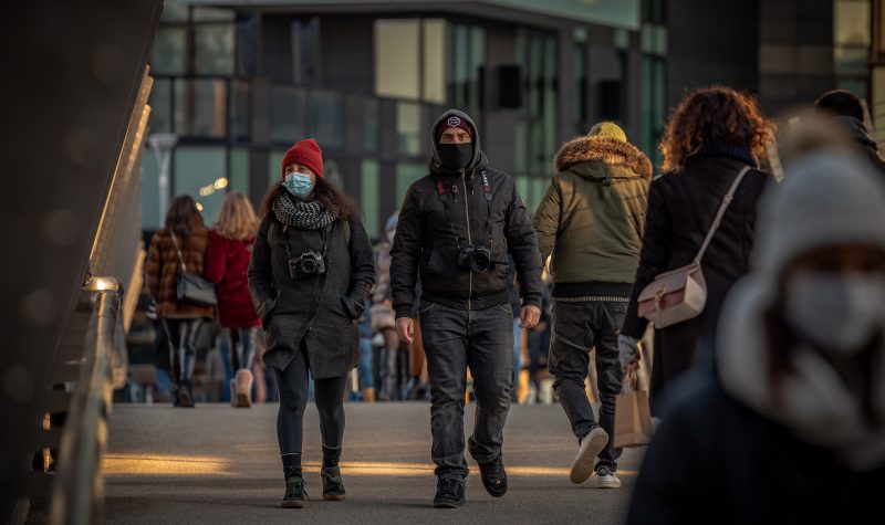 Un homme et une femme habillé en touristes avec appareil photos marchent dans la rue avec d'autres personnes, ils sont masqués.