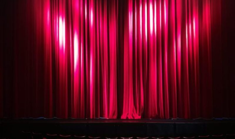 A red curtain is closed on stage with spotlights shining on it. There are rows of red theatre seats before the stage.