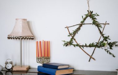 A series of items, including a Menorah, have been placed on a brown table. There is also a lamp, and a handmade Star of David hangs from the wall.