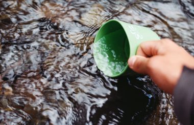 A hand sits a green coffe mug into a stream of water to fill it up.