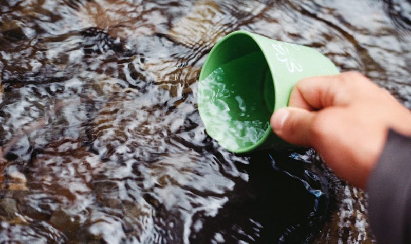 A hand sits a green coffe mug into a stream of water to fill it up.