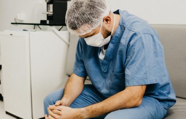 A person wearing blue medical scrubs, a protective mask, sits on a hospital bench in a white room, looking down at their feet