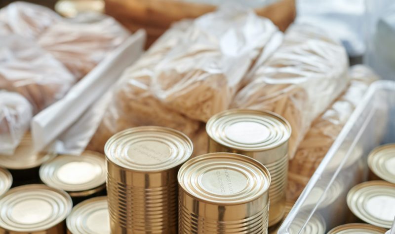 Stacks of non-perishable food items can be seen. There are piles of silver cans, plastic bags of dried food, and bottles of water.