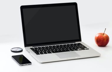 A laptop is seen on a white surface, with a phone placed to its left and an apple placed to its right.