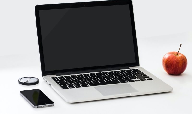 A laptop is seen on a white surface, with a phone placed to its left and an apple placed to its right.