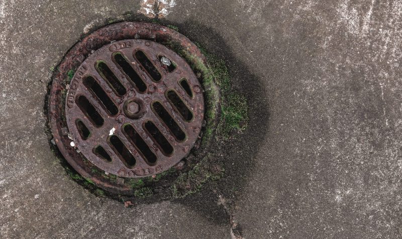 A round sewage drain is shown from above sitting on a paved street.