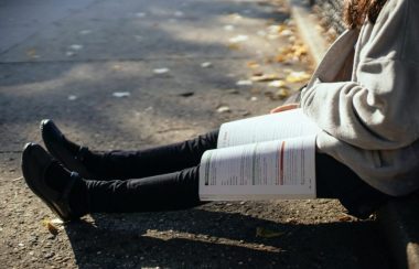 Les jambes d'une étudiante assise sur le trottoir. Un manuel de classe est ouvert sur les jambes.
