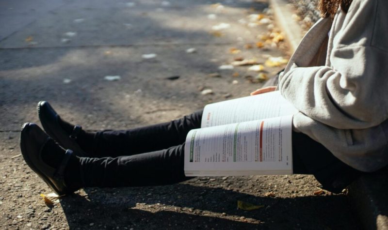 Les jambes d'une étudiante assise sur le trottoir. Un manuel de classe est ouvert sur les jambes.