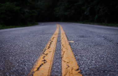 Asphalt Road pictured. Two solid painted yellow lines run down the middle of the road. Two lanes appear on the left and right of the yellow line. Trees sit along the sides of the road