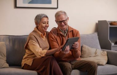 Un couple de personnes aînées assis sur un sofa regardent ensemble une tablette.