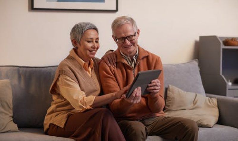 Un couple de personnes aînées assis sur un sofa regardent ensemble une tablette.