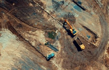 An overhead photo of a yellow excavator filling a dump truck with soil in a construction site.