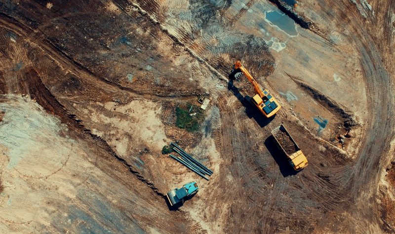 An overhead photo of a yellow excavator filling a dump truck with soil in a construction site.