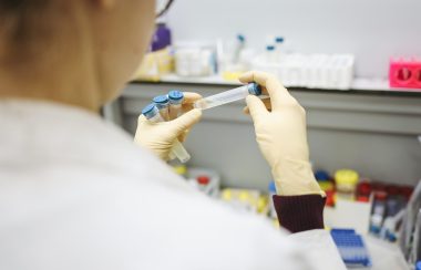 Someone wearing a whit lab coat is seen from behind, holding a number of unopened vials in their hands.