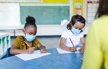 Two young children, wearing protective masks, sit side-by-side at a classroom table, writing on blank paper with coloured pencils. A chalkboard is seen in the background.