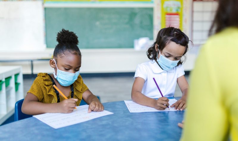 Two young children, wearing protective masks, sit side-by-side at a classroom table, writing on blank paper with coloured pencils. A chalkboard is seen in the background.