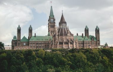 A view of the Parliament buildings is seen over a forested area on a cloudy day.