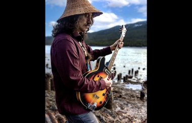 An indigenous musician wearing a cedar hat playing guitar outdoors beside a river.