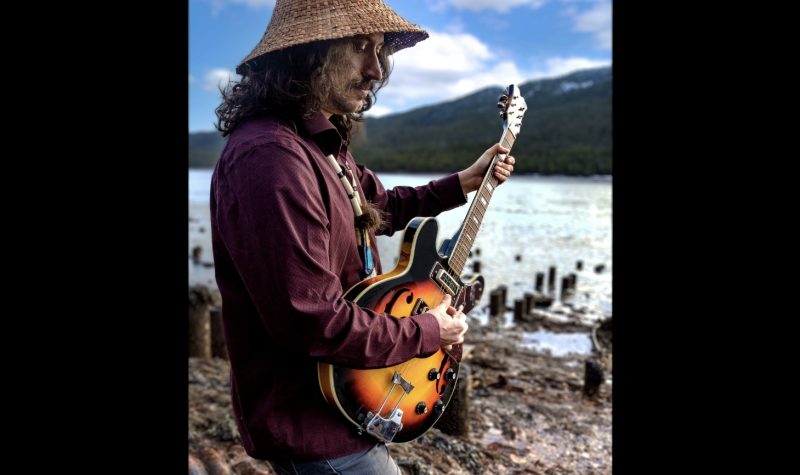 An indigenous musician wearing a cedar hat playing guitar outdoors beside a river.