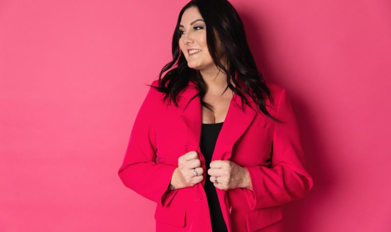 A brunette woman wearing a red blazer stands against a bright pink background.