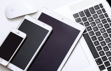 Un iPhone, un iPad, une tablette Samsung, une souris pour ordinateur Apple et un ordinateur portatif muni d’un clavier noir aux bordures blanches, écran fermé superposée les uns sur les autres aux dessus d’une table de travail de couleur blanche.