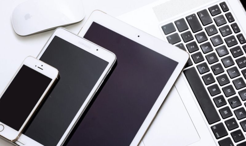 Un iPhone, un iPad, une tablette Samsung, une souris pour ordinateur Apple et un ordinateur portatif muni d’un clavier noir aux bordures blanches, écran fermé superposée les uns sur les autres aux dessus d’une table de travail de couleur blanche.