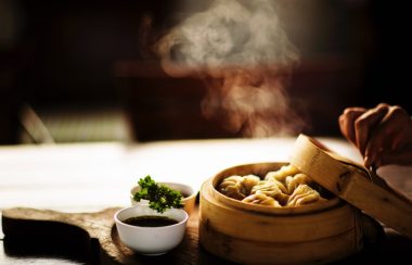 A hand is pulling the lid off a bamboo steam basket with dumplings inside while steam escapes.